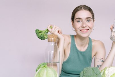 Woman in tank top smiling with broccoli, cabbage, and glass of water on table.